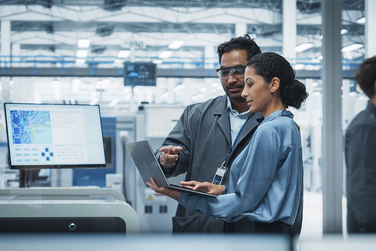 Two industrial engineers in lab coats and safety glasses reviewing operational data on a laptop inside a modern manufacturing facility.