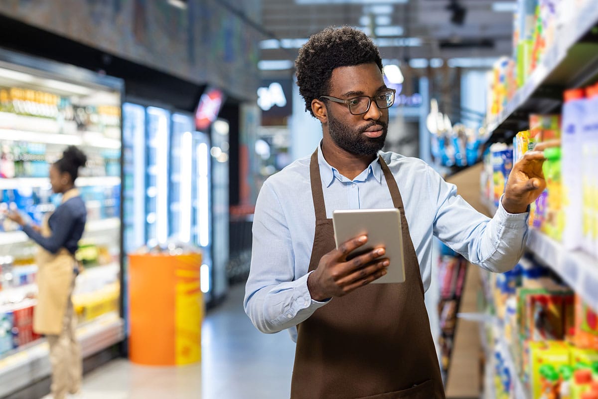 A retail worker in an apron holding a tablet while managing inventory and checking products on grocery store shelves.