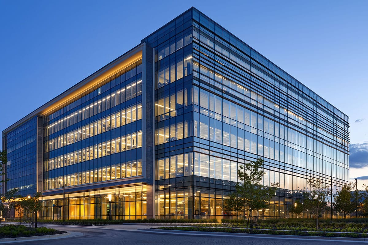 A modern multi-story glass corporate office building illuminated with warm interior lights at dusk.