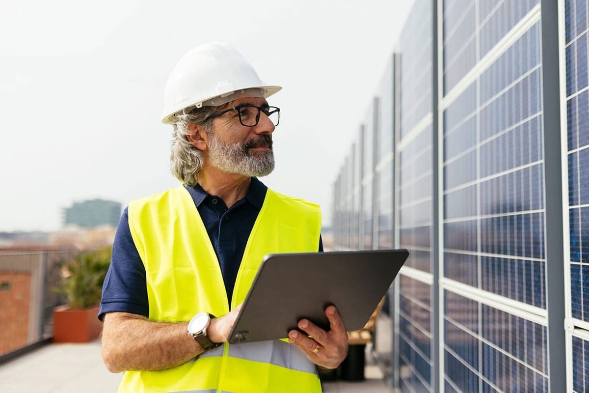 A utility worker wearing a white hard hat and yellow safety vest using a tablet in front of a large solar panel installation.