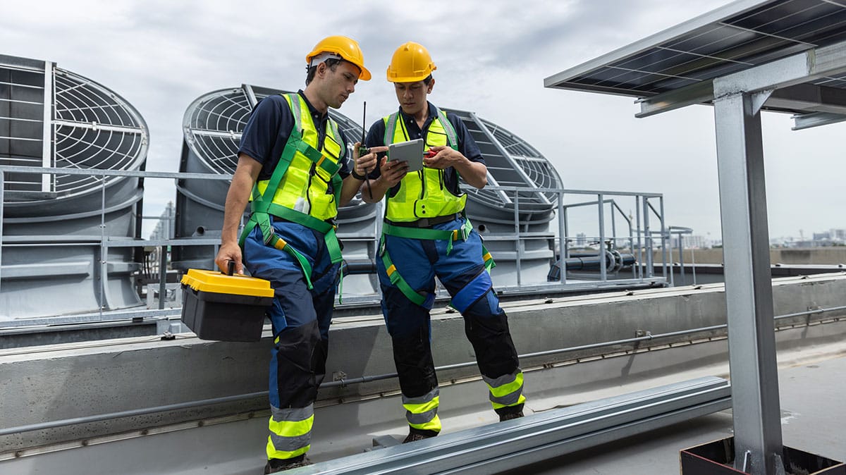 Rooftop workers in smart helmets and vests using a 5G tablet near large industrial ventilation units.